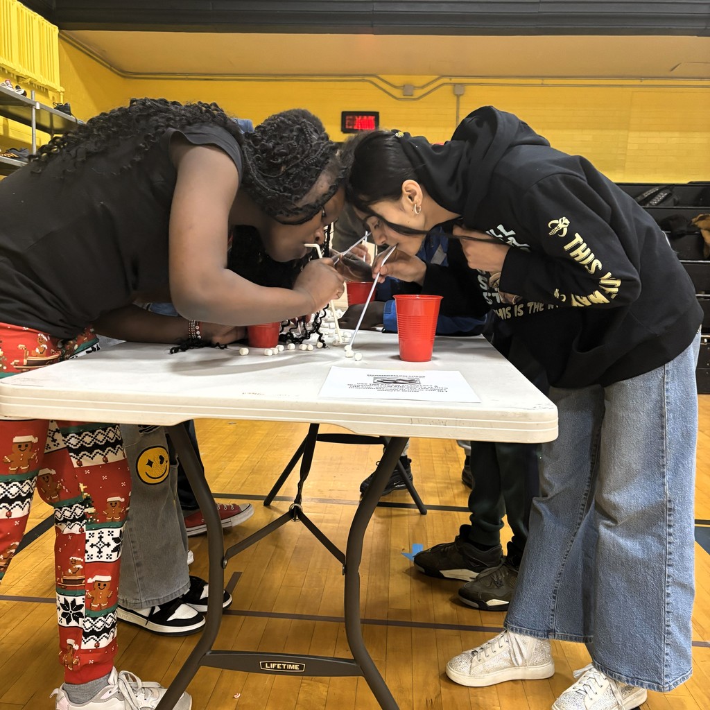 Students lean over a table, using straws to move marshmallows in a game.