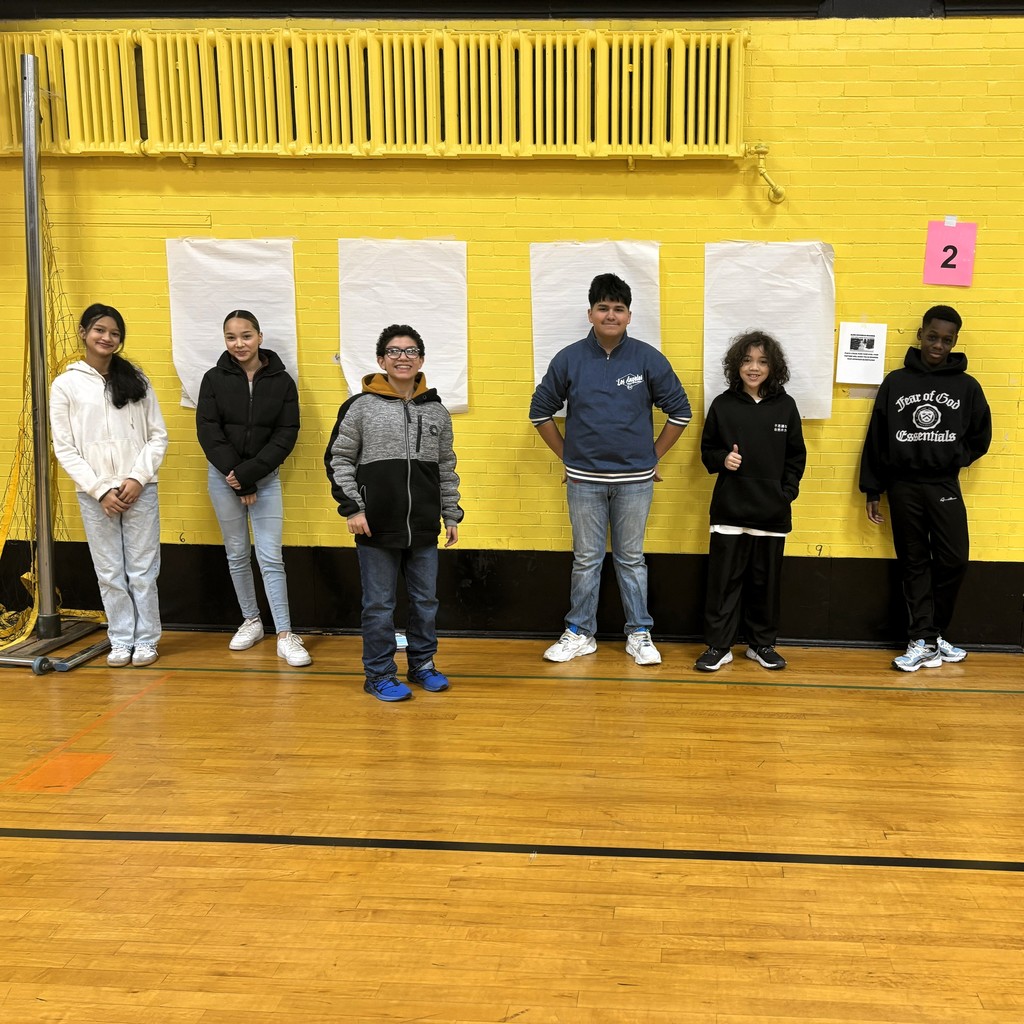 Six students stand in a line against a bright yellow brick wall in a gymnasium.