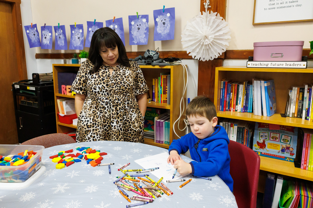 A teacher observes as a young student draws on a piece of paper.
