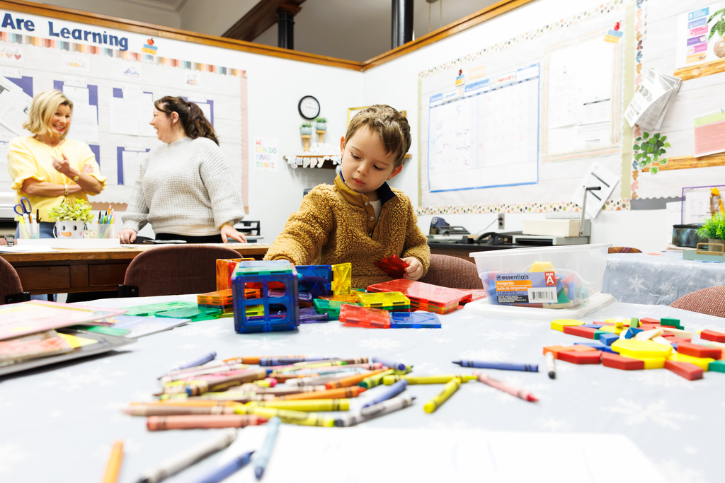 A child plays with building blocks while his caregiver and a teacher talk in the background.
