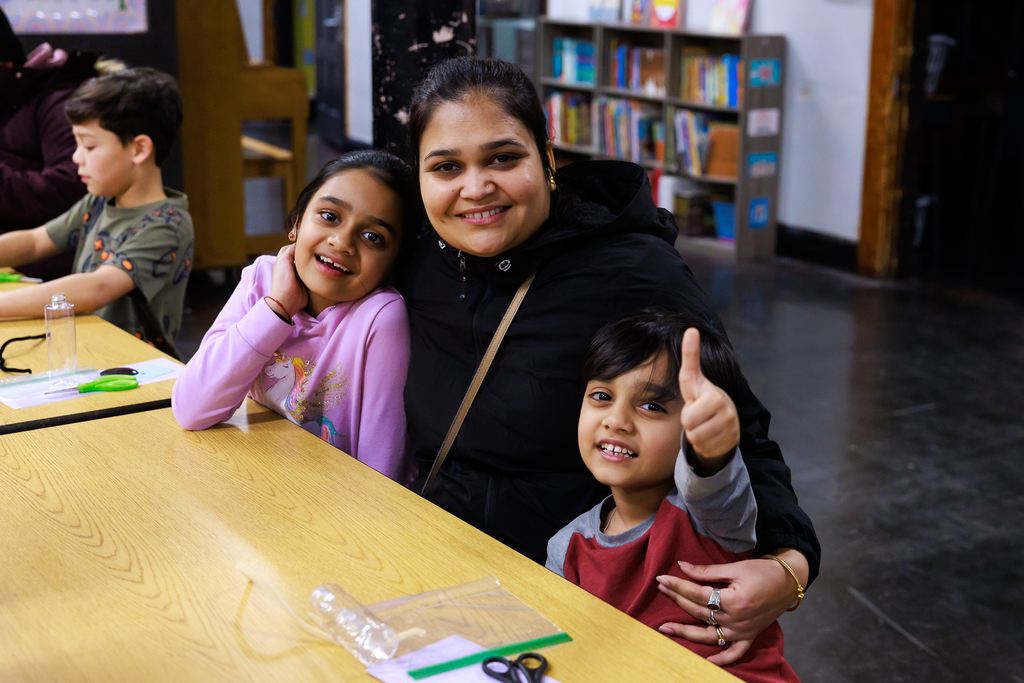 A woman smiles with two children, a girl in a pink unicorn shirt and a boy giving a thumbs up.