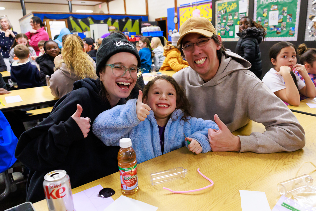 Two adults and one young child sit together and give the thumbs up, each one smiling.