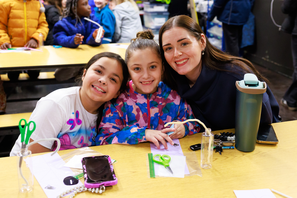 A woman and two young girls smile at the camera while sitting at a wooden table.