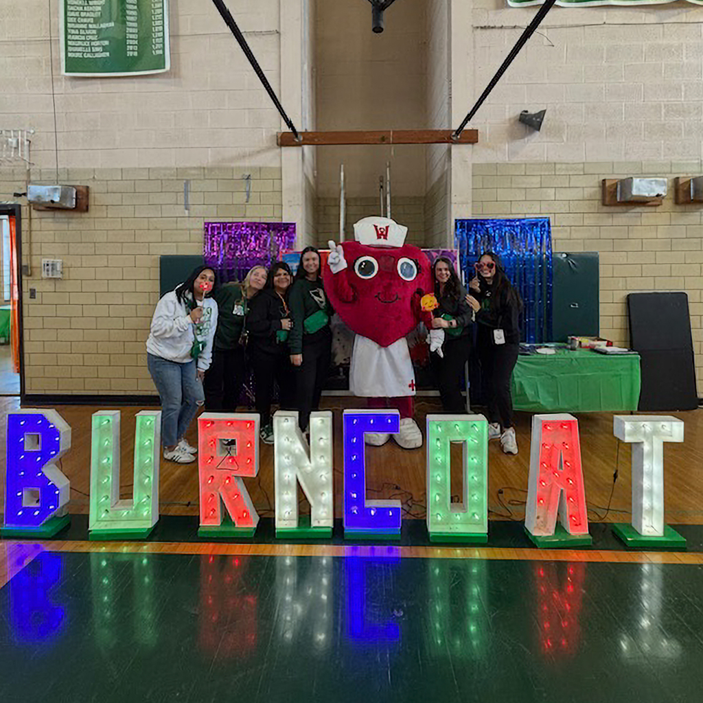 A group of people pose with a heart mascot in a gymnasium, in front of large illuminated letters spelling 'Burncoat'.