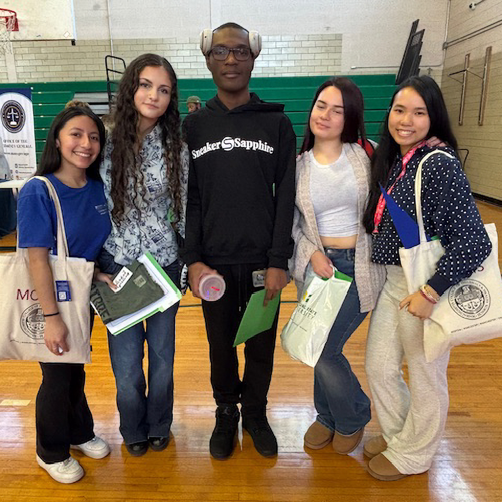 Five young people stand together in a gymnasium, holding bags and folders.
