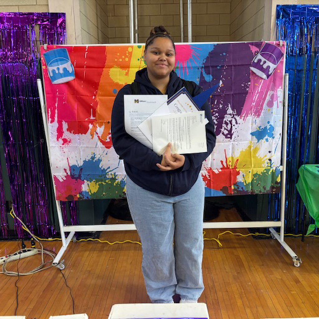 A young woman smiles, holding certificates and papers in front of a colorful paint splatter backdrop.
