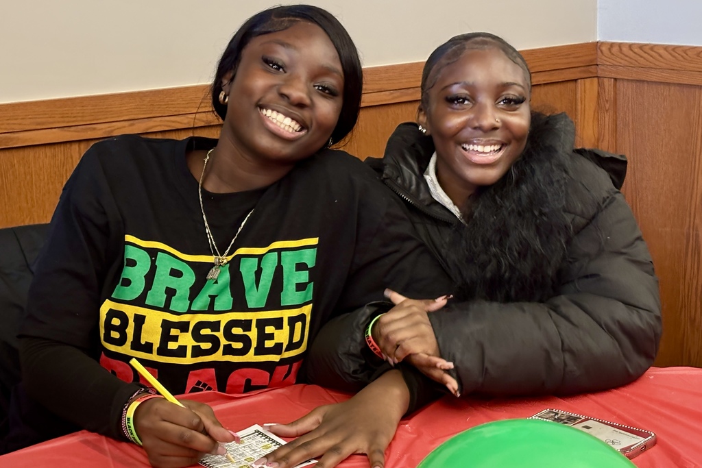 Two young Black women smile while sitting at a table, one wearing a shirt that says "BRAVE BLESSED BLACK".
