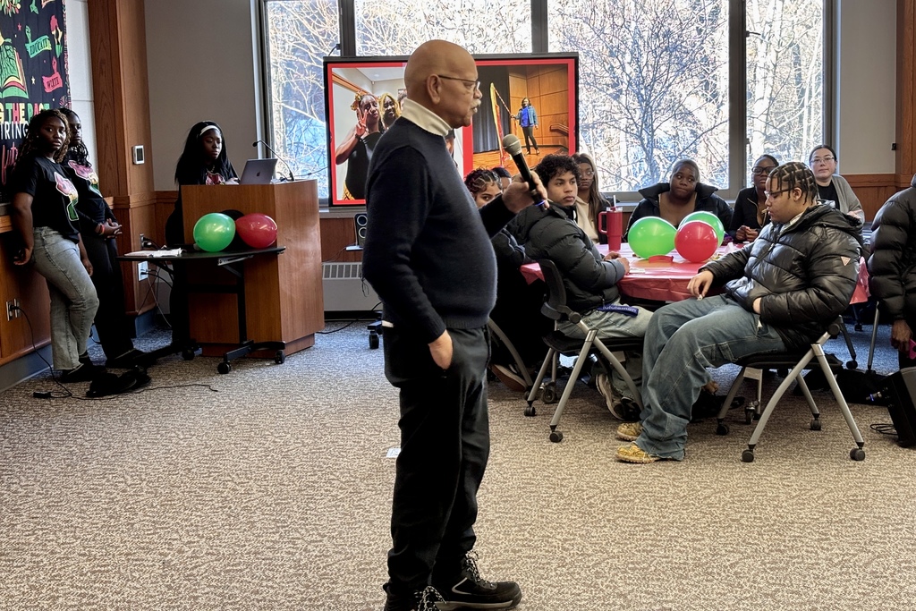 A speaker addresses a group of students seated at tables, with balloons and a presentation screen in the background.