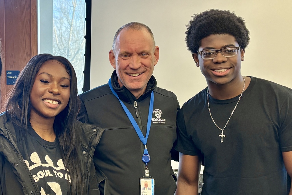 A smiling man in a black jacket with a Worcester Public Schools logo stands between two smiling young people.