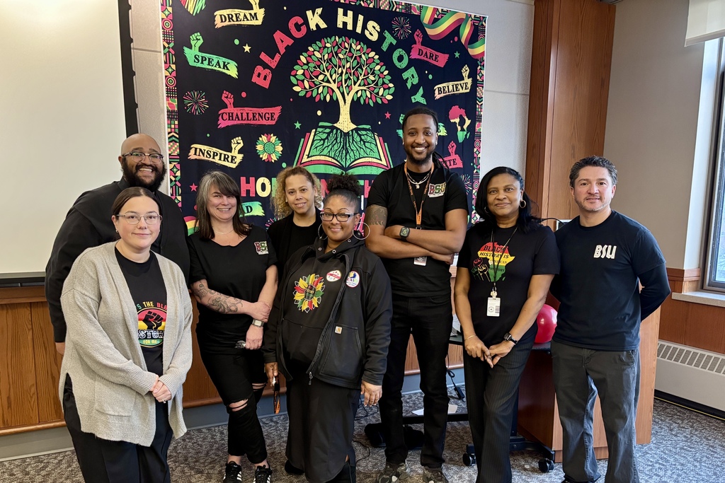 A diverse group of people stand together in front of a vibrant Black History Month banner.