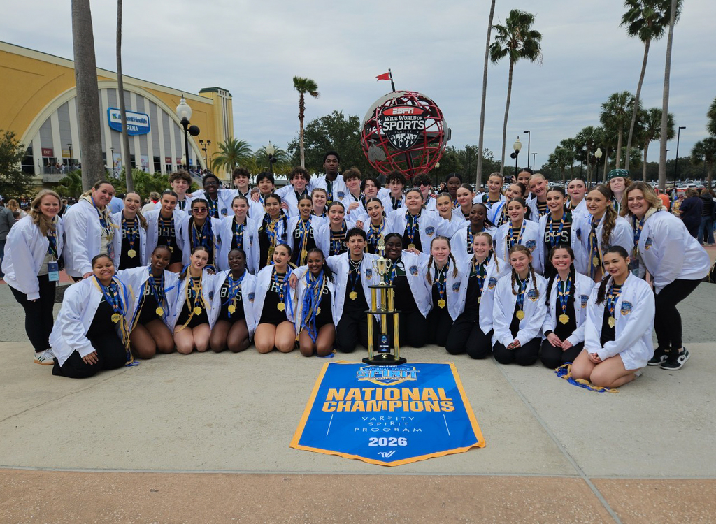 The Burncoat Spirit Team poses for a photo behind a National Championship trophy, and a banner that reads National Champions Varsity Spirit Program 2026. They are standing in front of a fountain and a statue that says "ESPN Wide World of Sports Complex."