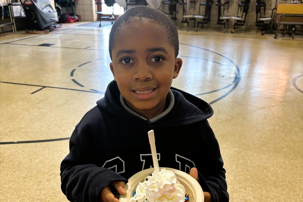 A young boy in a dark hoodie smiles while holding a bowl of whipped cream and sprinkles.