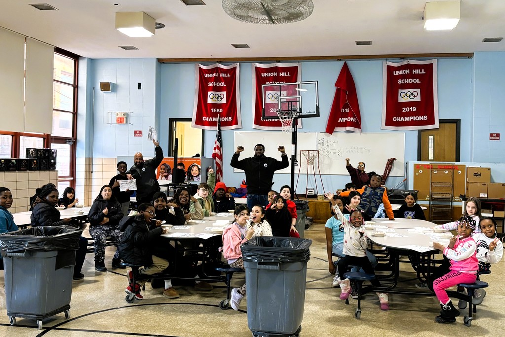 Students and adults gather in a school gymnasium with banners celebrating past championships.