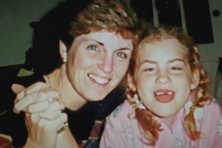 A woman with short brown hair and blue eyes smiles warmly next to a young girl with blonde pigtails.