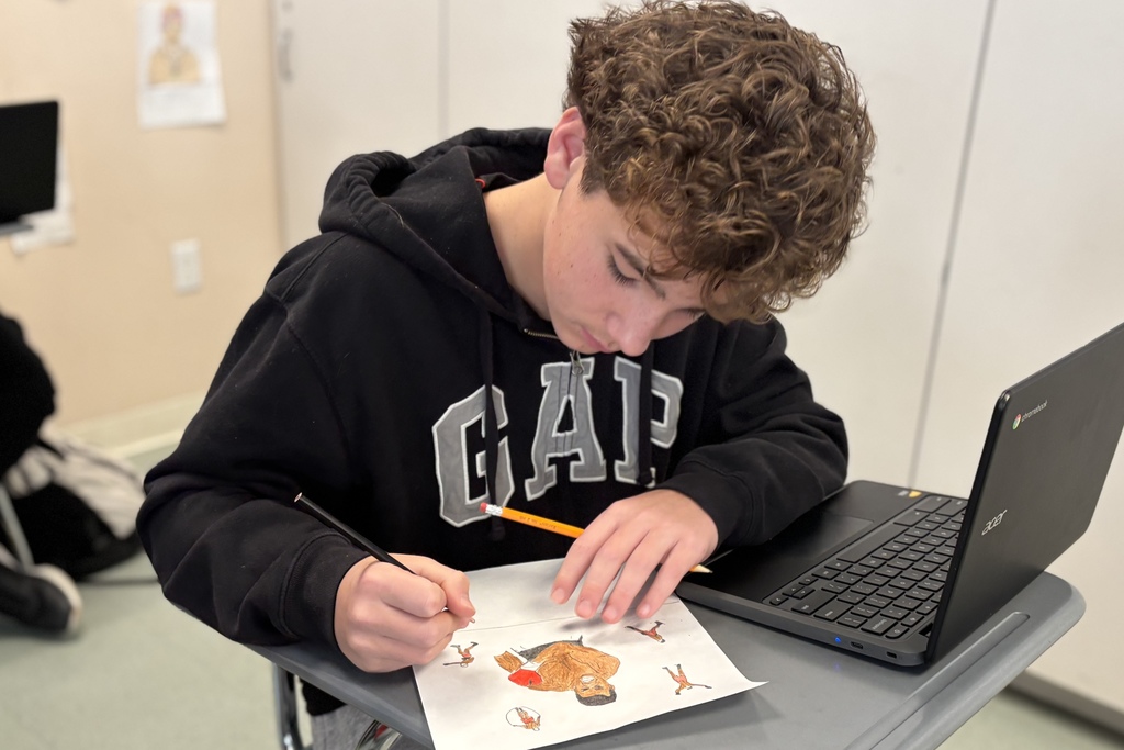 A young person with curly hair draws on a piece of paper while a laptop sits open on their desk.