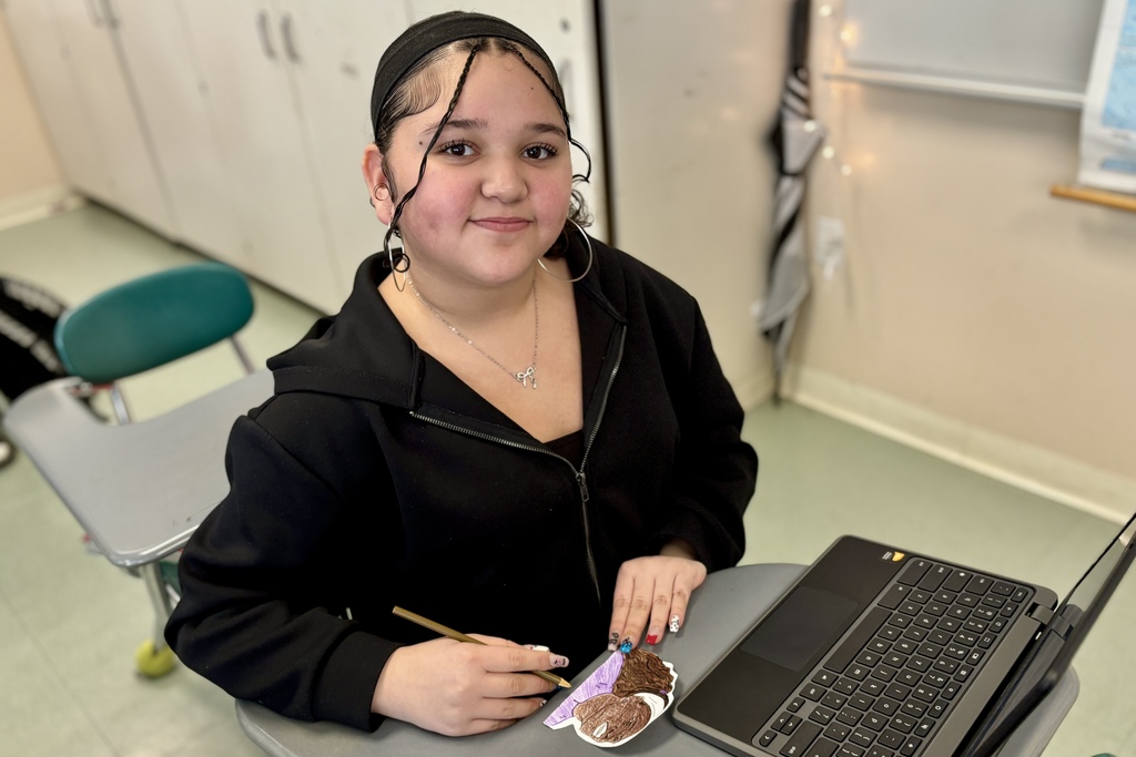 A young person with braided hair sits at a desk with a laptop and art supplies.