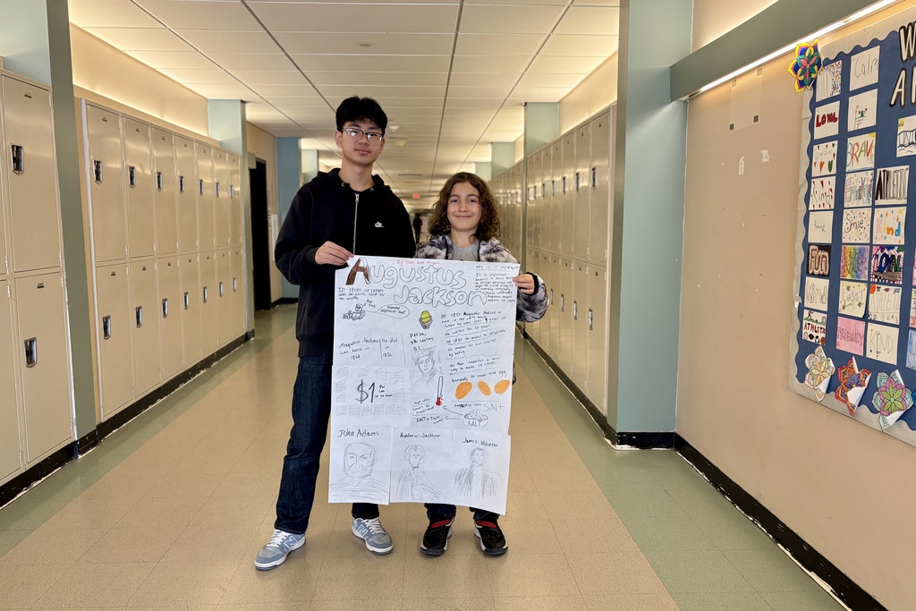 Two students stand in a school hallway holding a poster about Augustus Jackson, with lockers lining the walls.