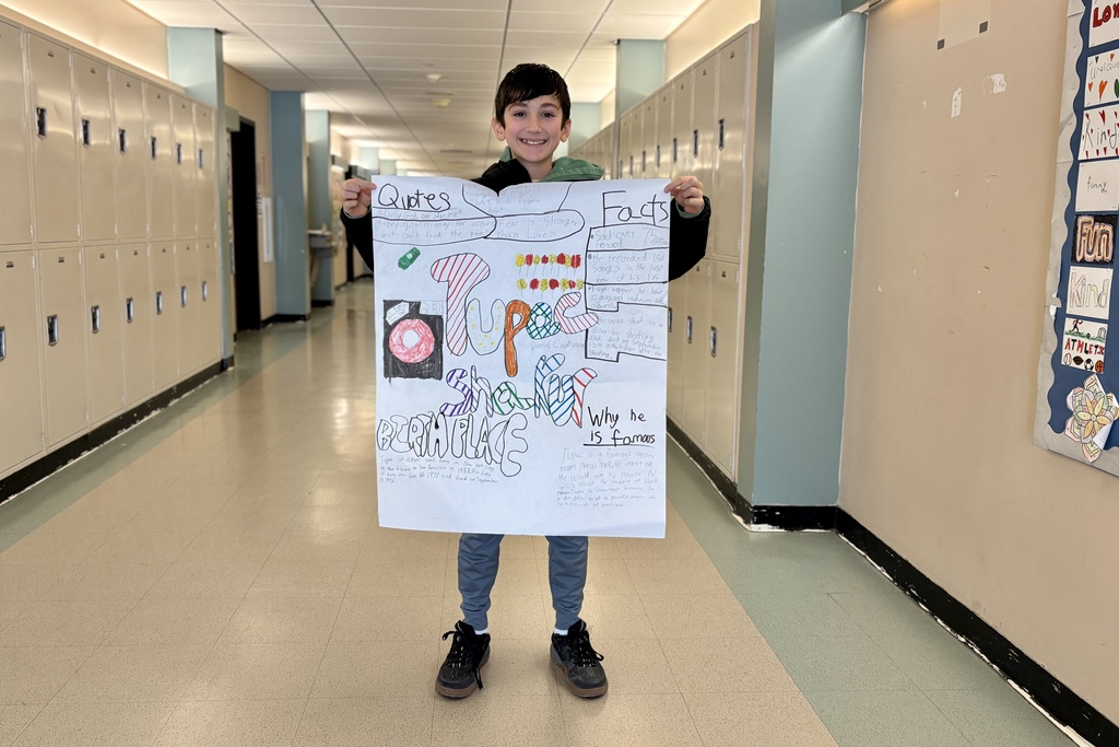 A young boy smiles while holding a poster about Tupac Shakur in a school hallway with lockers.
