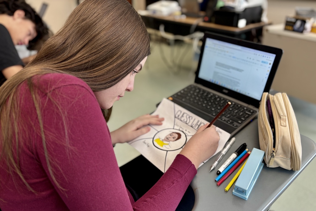 A student works on a 'Guess Who' drawing at a desk with a laptop and art supplies.