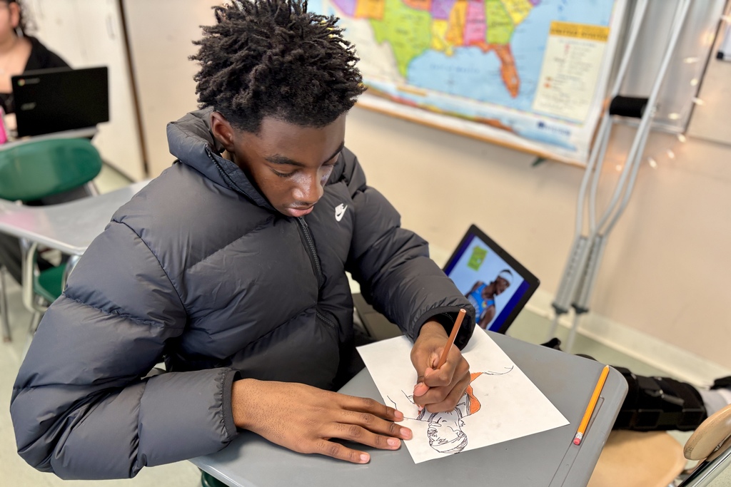 A young person in a puffer jacket draws on a piece of paper at a desk in a classroom.