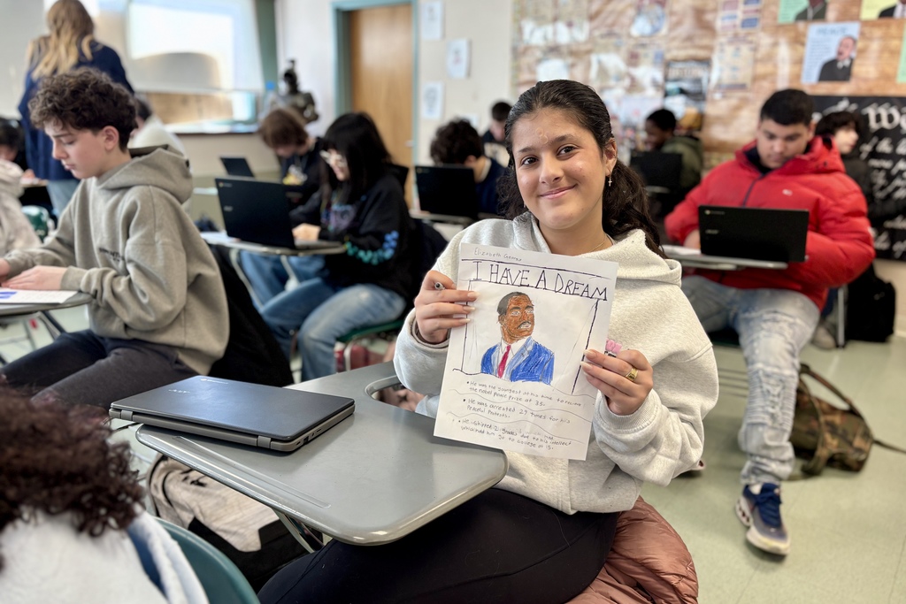 A student smiles while holding a drawing and paper titled 'I HAVE A DREAM' in a classroom.
