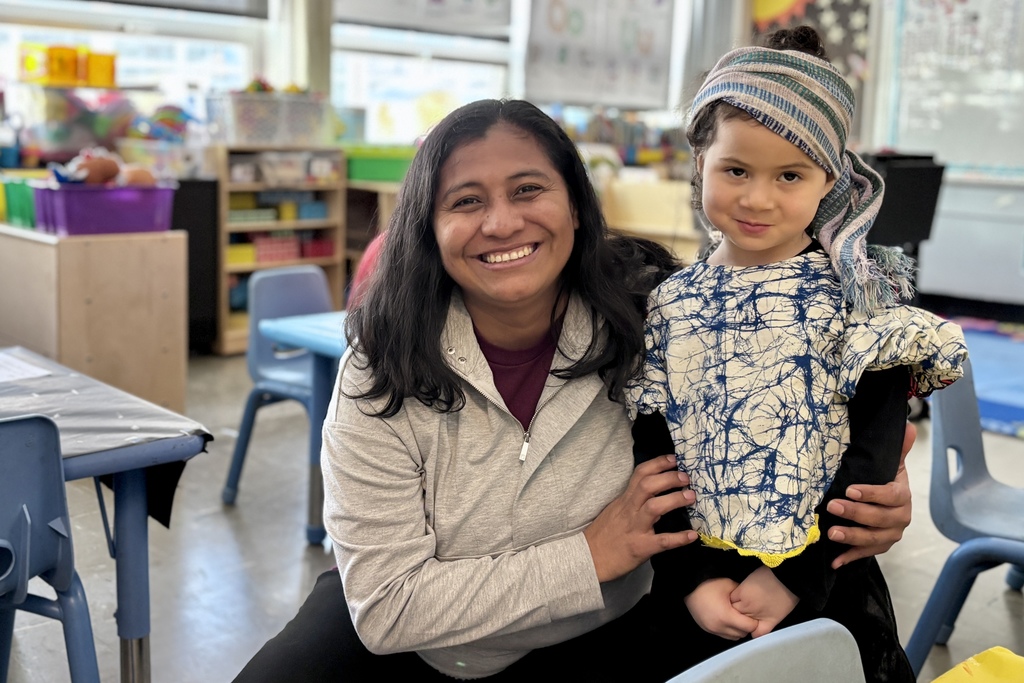 A smiling woman and a young child pose together in a classroom setting.