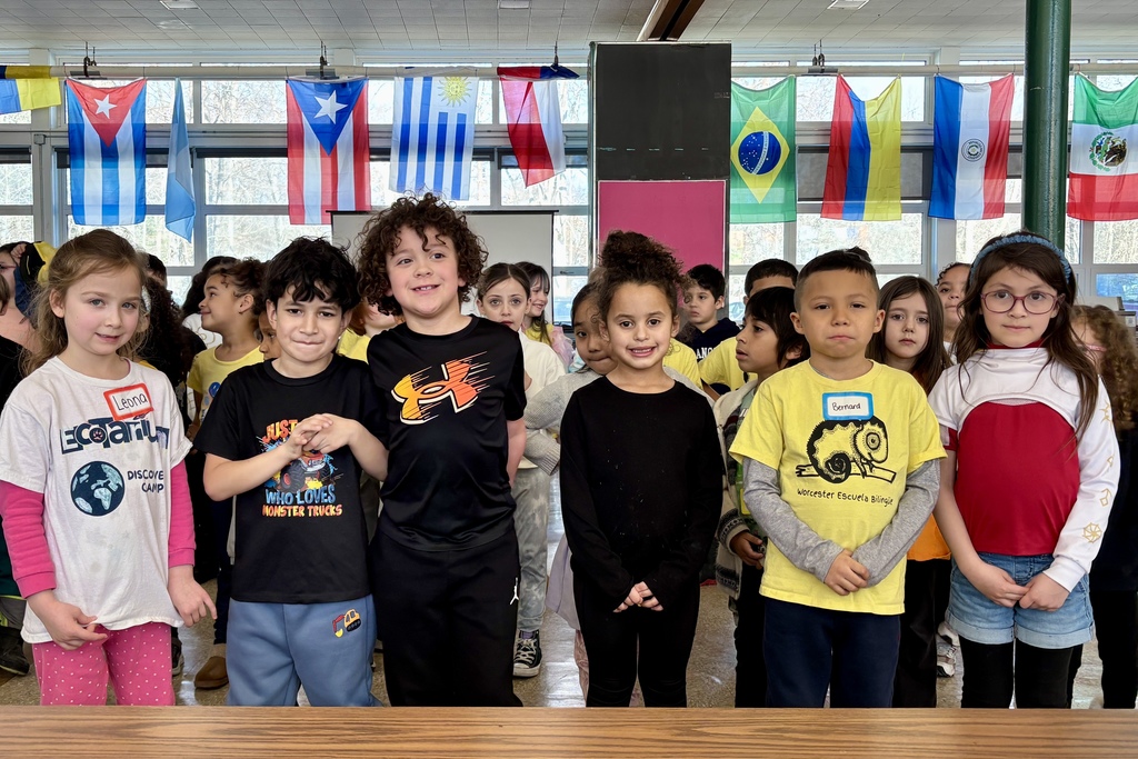 A diverse group of children stand in front of flags from various countries, including Cuba, Puerto Rico, and Brazil.
