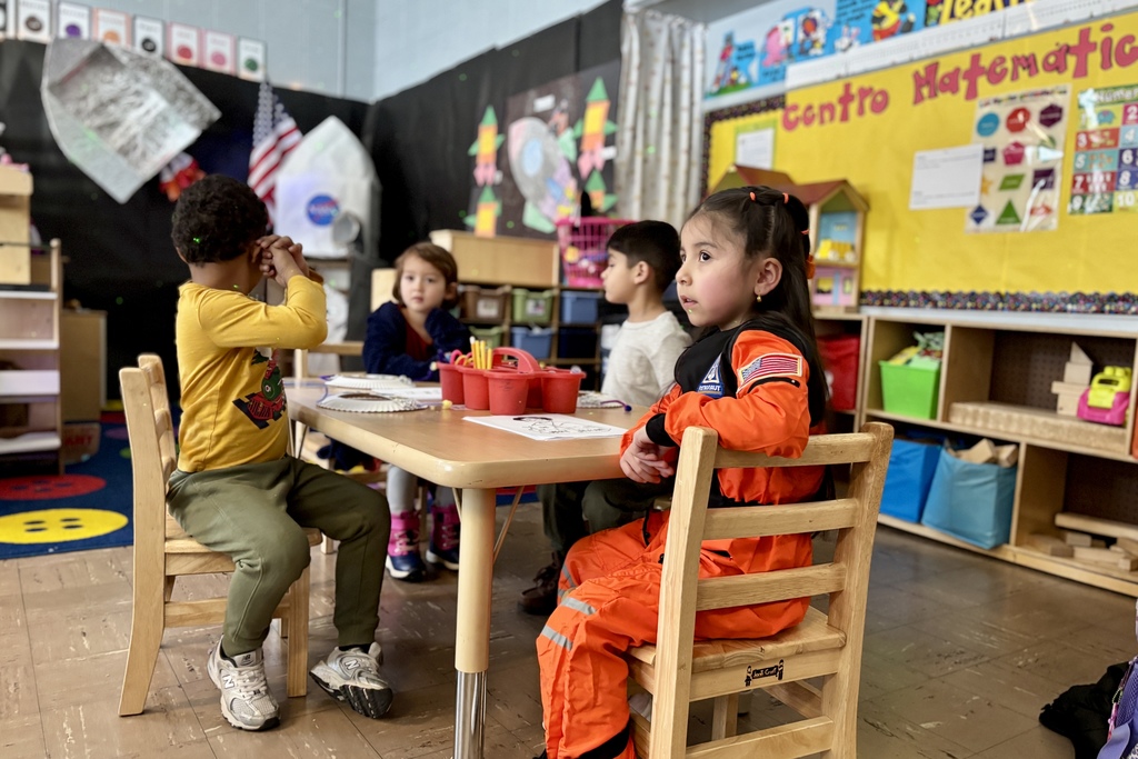 Children sit at a table in a classroom decorated with space-themed artwork and a NASA rocket.