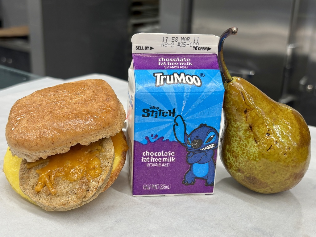 A breakfast sandwich with cheese, a carton of TruMoo chocolate milk featuring Stitch, and a pear are arranged on a white surface.