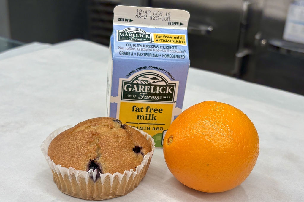 A blueberry muffin, an orange, and a carton of Garelick Farms fat-free milk are arranged on a white surface.