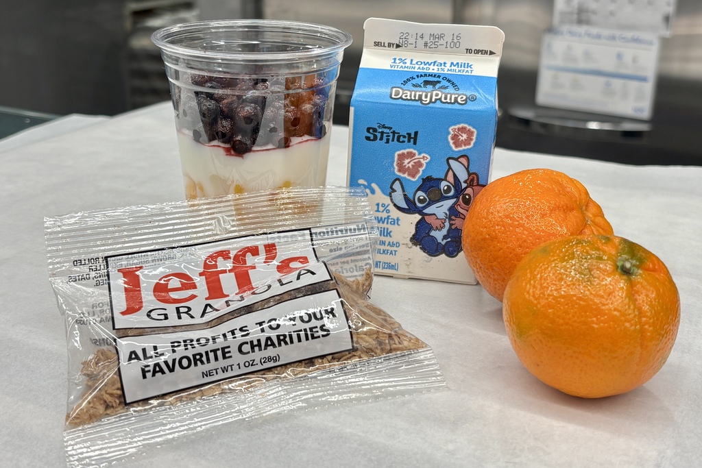 A breakfast spread includes a yogurt parfait with berries, two clementines, a small carton of DairyPure milk featuring Stitch, and a packet of Jeff's Granola.