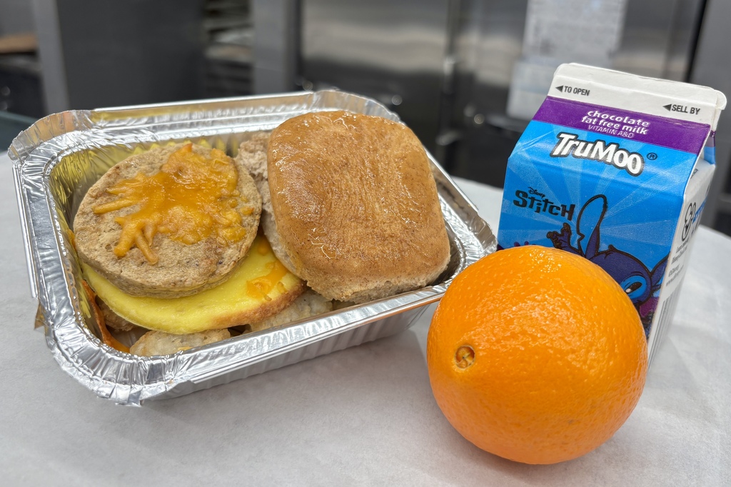 A foil container holds a breakfast sandwich with melted cheese, next to an orange and a carton of TruMoo chocolate milk featuring Disney's Stitch.