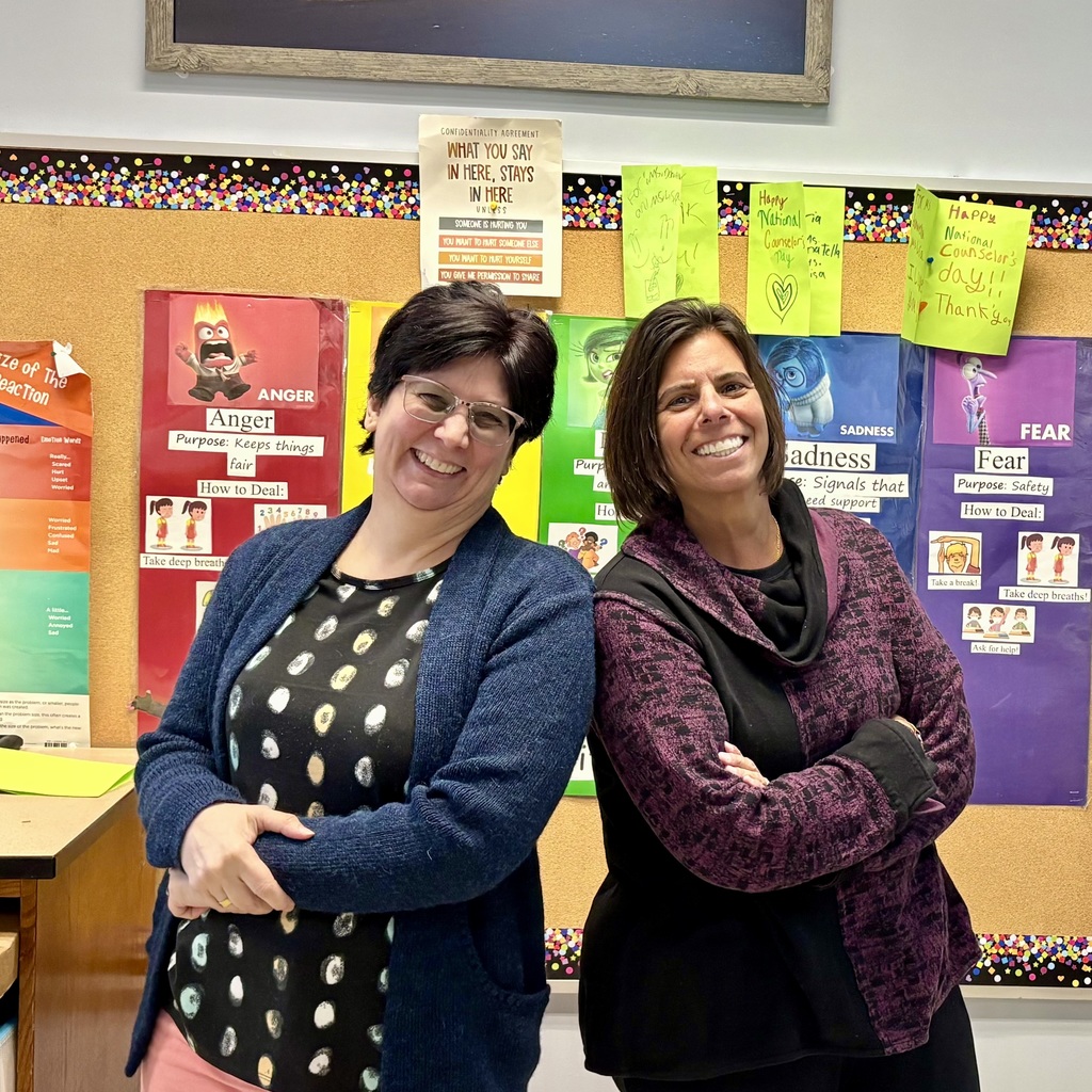 Two smiling women stand back-to-back in front of a bulletin board decorated with colorful confetti and posters about emotions.