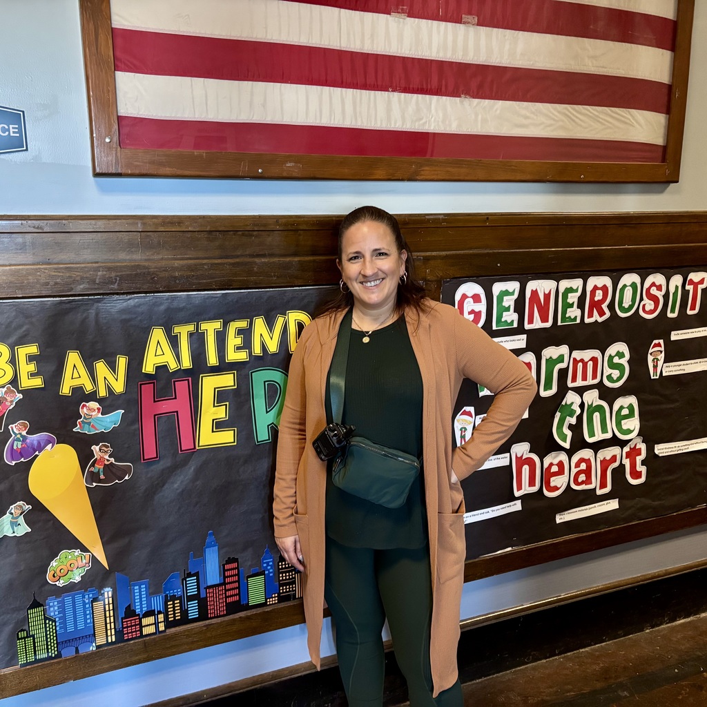A woman smiles in front of a bulletin board with superhero cutouts and the words 'BE AN ATTEND HERO'.