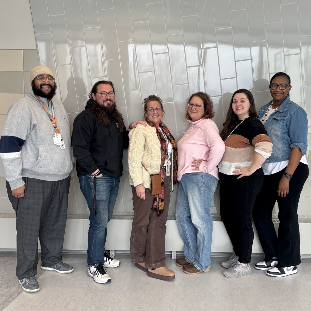 Six people stand together in front of a tiled wall, smiling.