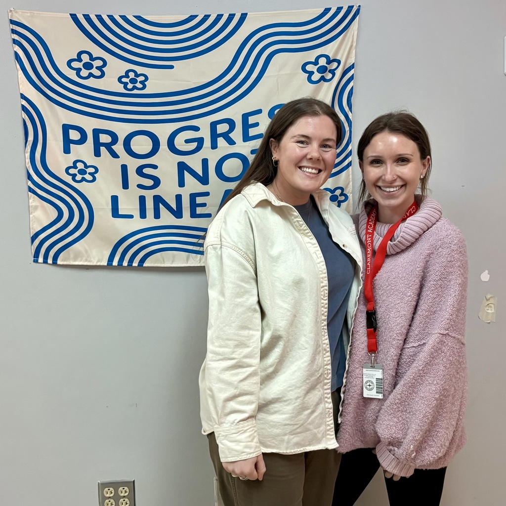 Two smiling women stand in front of a banner that reads "PROGRESS IS NOT LINEAR" with blue wavy lines and flowers.