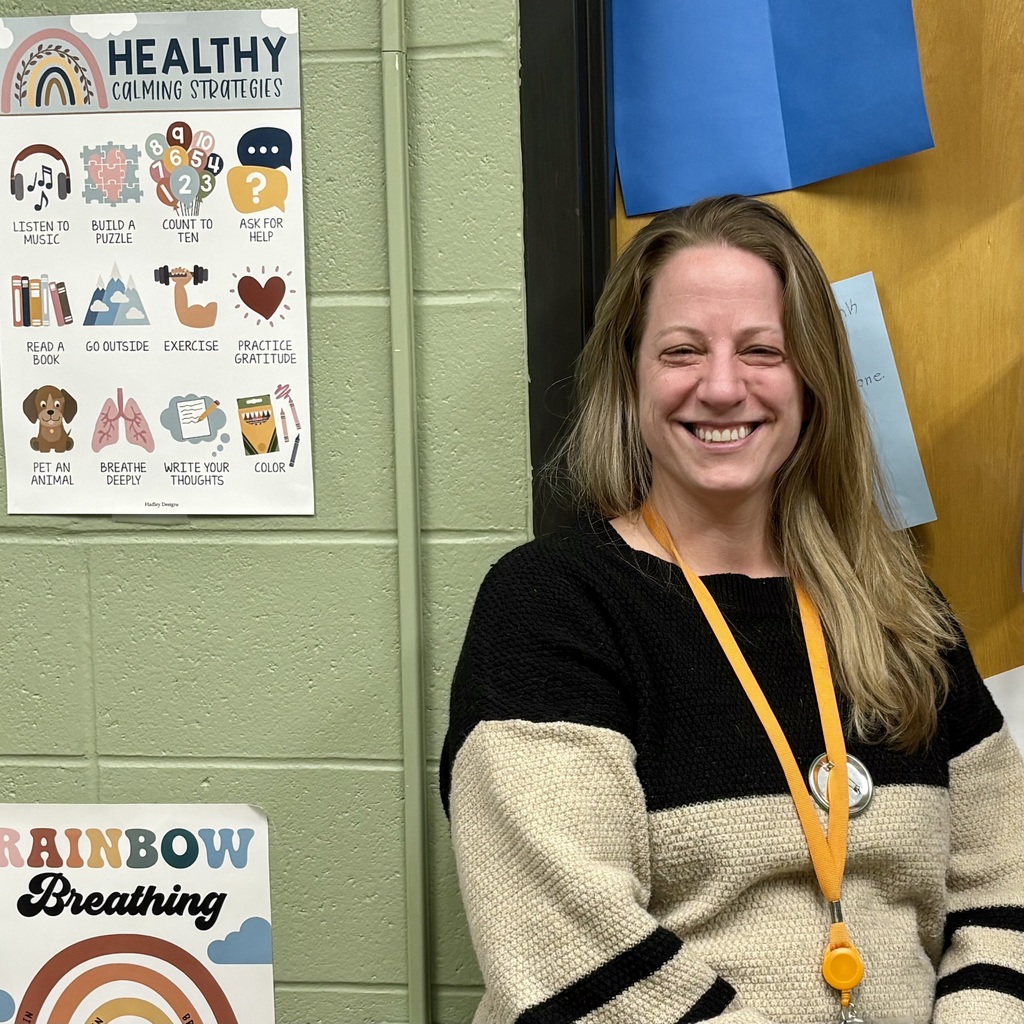 A woman smiles broadly in front of a wall decorated with "Healthy Calming Strategies" and "Rainbow Breathing" posters.