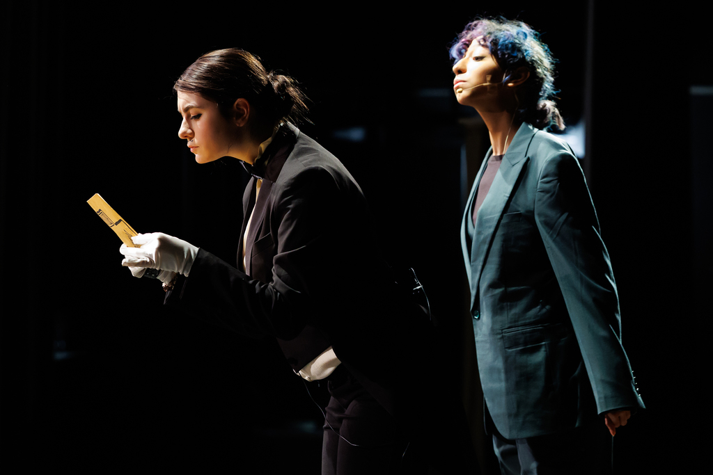 A person in a black suit and white gloves holds a golden ticket, looking down intently.