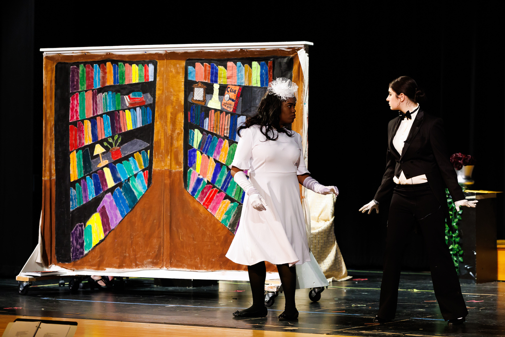 Two actors stand on a stage in front of a colorful, painted bookshelf backdrop.