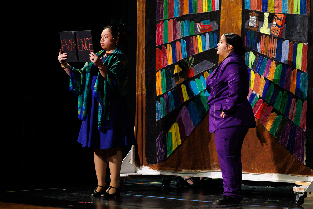 Two actors stand on a stage in front of a colorful, painted bookshelf backdrop, one is holding a book with the words 'evidence'.