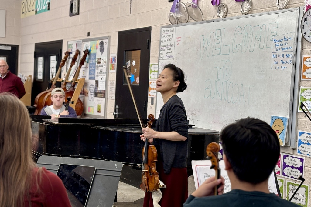 A music instructor leads a violin lesson in a classroom with a whiteboard that says 'WELCOME TO BURNCOAT'.
