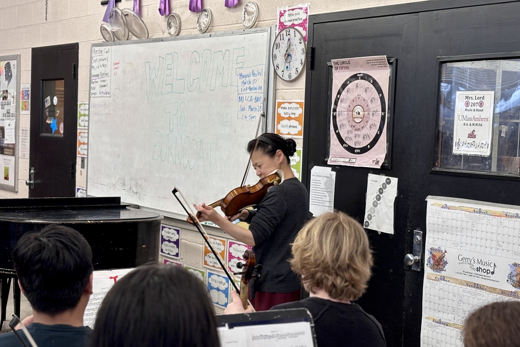 A woman plays the violin in a classroom with students in the foreground, a piano to the left, and educational posters on the wall.