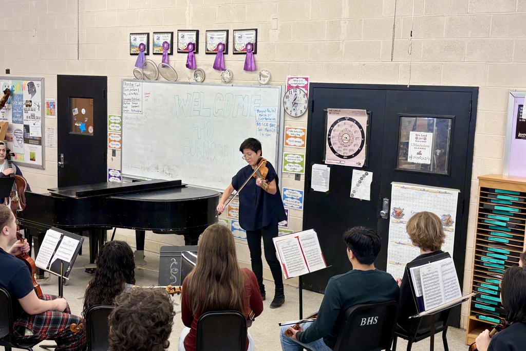A music instructor plays the violin for a class of students seated with their instruments.