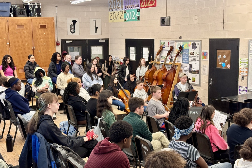 Students and adults are seated in chairs, facing forward, in a room with musical instruments and posters on the walls.