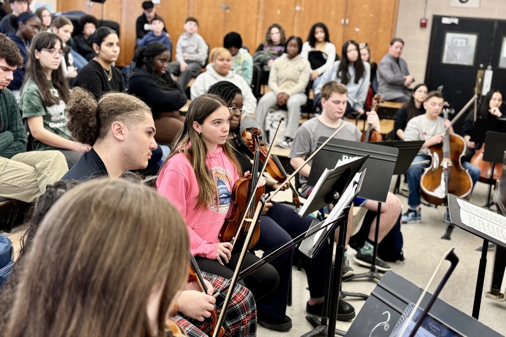 A diverse group of young musicians practice in an orchestra setting, holding violins and cellos.