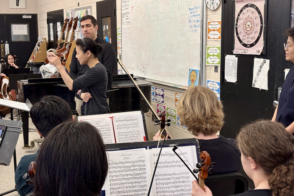 A music instructor gestures while students with violins practice in a classroom with sheet music.