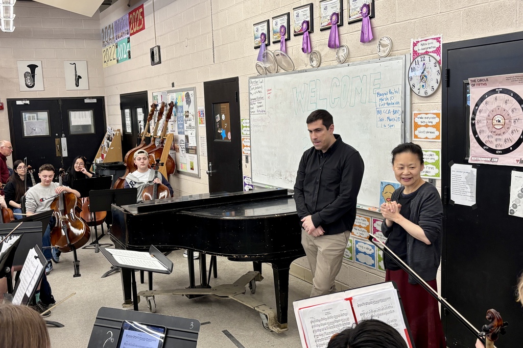 A music class with students playing cellos and a piano in the foreground, with instructors standing near a whiteboard.