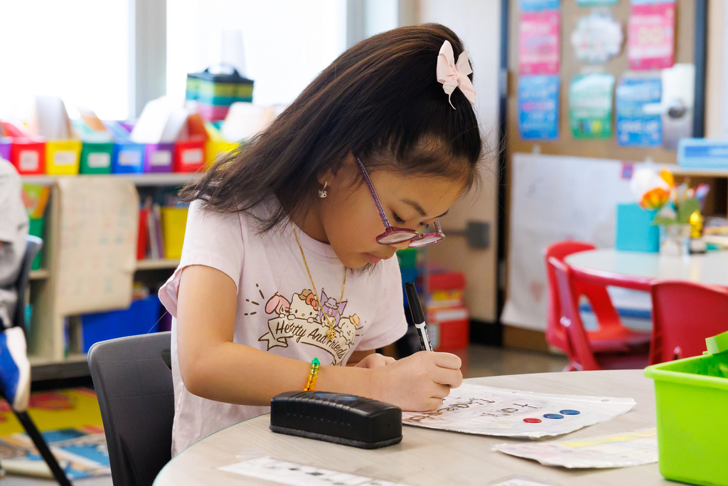 A young girl with a pink bow in her hair focuses on writing at a classroom table.