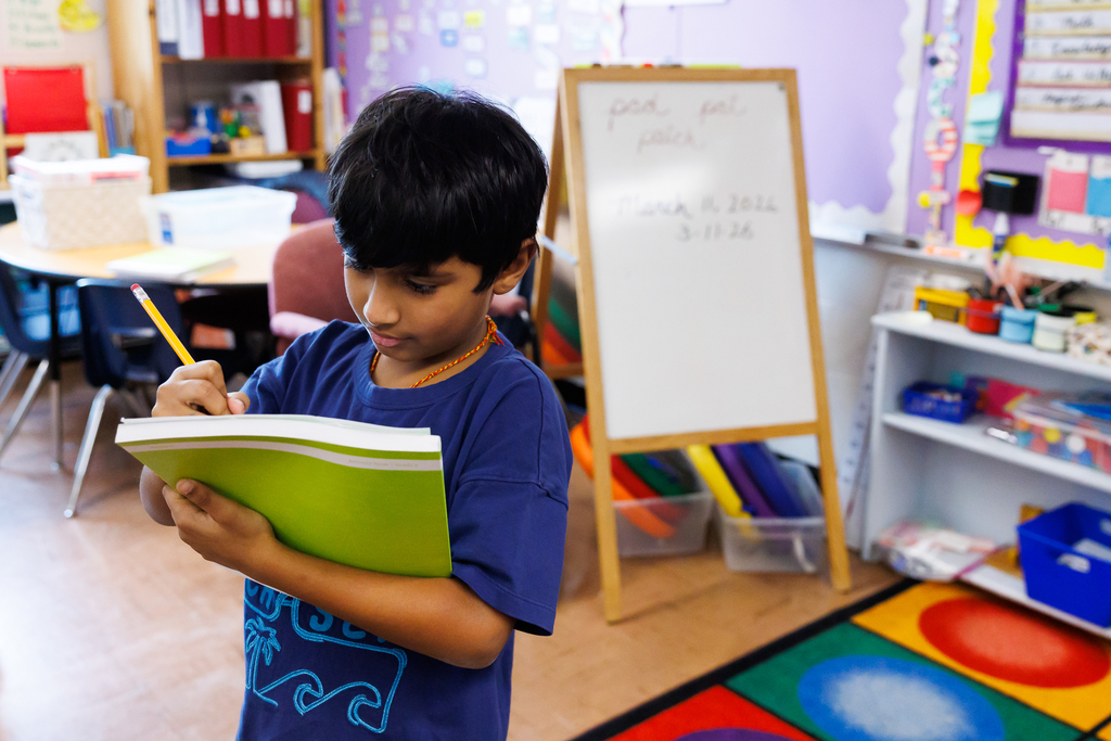 A young boy in a blue t-shirt writes in a green notebook with a yellow pencil in a classroom.