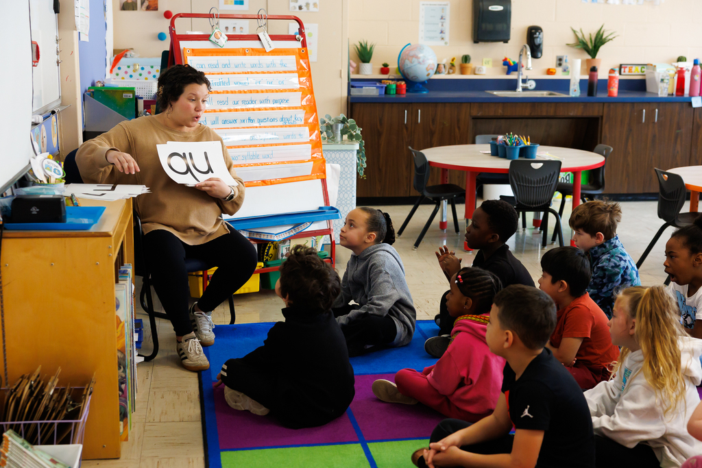 A teacher holds up a card with 'qu' to a group of young students sitting on a colorful rug in a classroom.